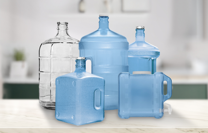 Five large water jugs, some clear and some blue, in various shapes and sizes are arranged on a marble countertop with a blurred kitchen background.