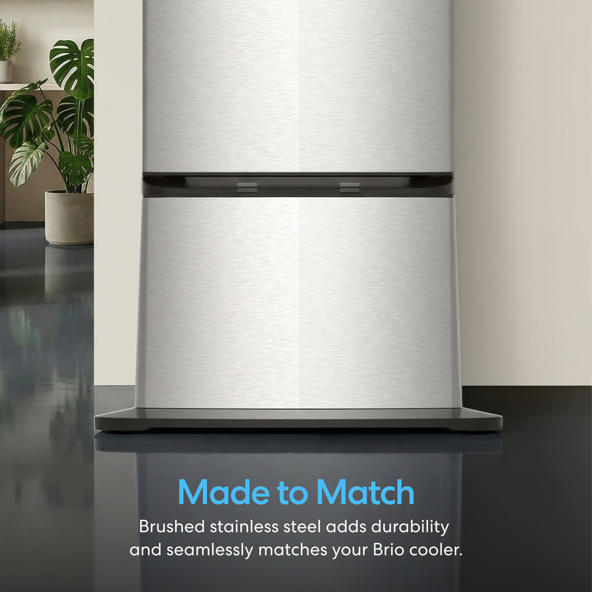 A close-up of a brushed stainless steel Cooler Base for Water Dispenser - 745 Series, with text reading: Made to Match—Brushed stainless steel adds durability and pairs seamlessly with your Brio 745 Series cooler. A plant and chair appear in the background.
