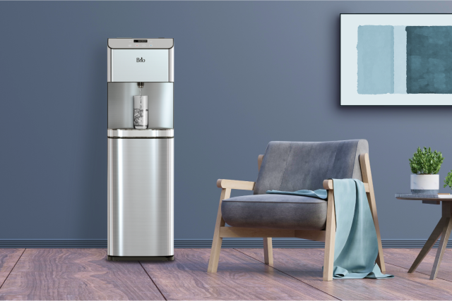 A modern water dispenser with a glass of water is next to a gray armchair with a blue throw blanket, a small plant on a table, and a framed abstract painting on a gray wall.