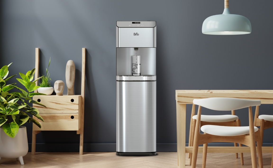 A modern stainless steel water dispenser stands against a dark gray wall, with a glass filling up. Nearby are a wooden cabinet with potted plants and a wooden dining table with chairs under a blue hanging lamp.