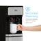 A hand uses a large push-paddle on the Stainless Steel Bottom Load Water Cooler - 300 Series to fill a white mug. Steam rises from the mug. Text reads: No-Contact Dispensing—large paddles ensure sanitary use.
