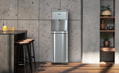 Modern kitchen or bar area with a Brio stainless steel bottom-loading water cooler centered against a gray textured wall. To the left is a counter with three wooden bar stools and a glass pitcher with orange juice, and to the right is a built-in shelf with decorative vases and potted plants.