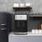 A 3-Stage Stainless Steel Bottleless Countertop Water Dispenser - 300 Series sits on a kitchen counter between a refrigerator and shelves with dishes. Three canisters are on the counter, and the wall has a gray hexagon tile backsplash. This sleek dispenser features self-cleaning technology for convenient maintenance and ensures pure, fresh water every time.