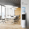 A modern office featuring white desks, chairs, expansive windows, and the White Top Load Water Cooler - 320 Sim Series with a NEW Slim Design prominently displayed in the foreground. The bright space is accented by wood finishes and stylish lighting.