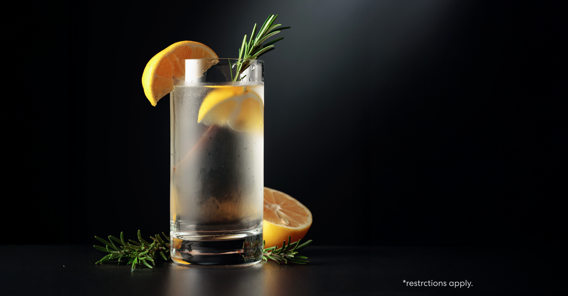 A tall glass of sparkling water garnished with lemon slices and a sprig of rosemary sits on a dark surface, with additional rosemary and a sliced lemon beside it. The drink glows against a dramatic black background, emphasizing its refreshing and crisp appearance.