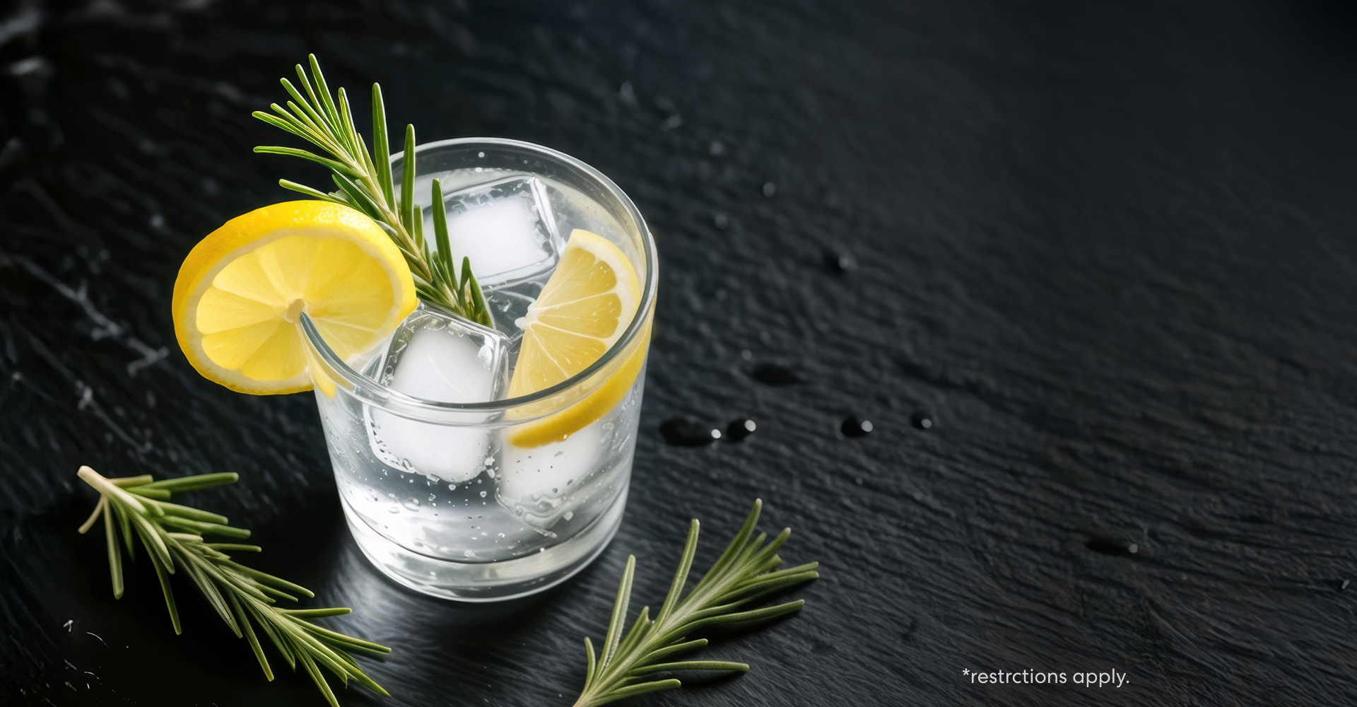 Glass of sparkling water with ice cubes, lemon slices, and rosemary sprigs on a dark stone surface, highlighting freshness and hydration. Text in corner reads “restrictions apply.”