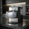 A Countertop Nugget Ice Maker - 740i in stainless steel and black, featuring a clear bin filled with nugget ice, sits in a modern kitchen with dark cabinets and gray countertops.
