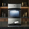 The Countertop Nugget Ice Maker - 740i, crafted from stainless steel, sits on a dark marble countertop overflowing with fresh nugget ice. A metal ice scoop rests in front of the machine. Behind it, wood paneling, shelves stocked with glassware, and golden bar accessories complete the background.