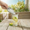 A hand pours ice cubes—freshly made with the Countertop Nugget Ice Maker - 740i—from a metal scoop into a glass of lemonade with lemon slices and mint, splashing water onto a wooden table outdoors with greenery in the background.