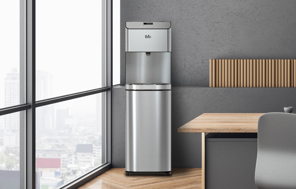 A modern silver water dispenser stands next to a wooden desk and gray chair in a contemporary office with large windows overlooking a cityscape.
