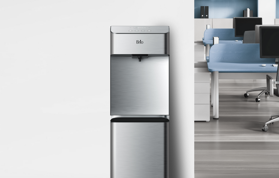 A stainless steel water cooler stands against a white wall in an office; blue chairs and desks are visible in the background, suggesting a modern workplace environment.