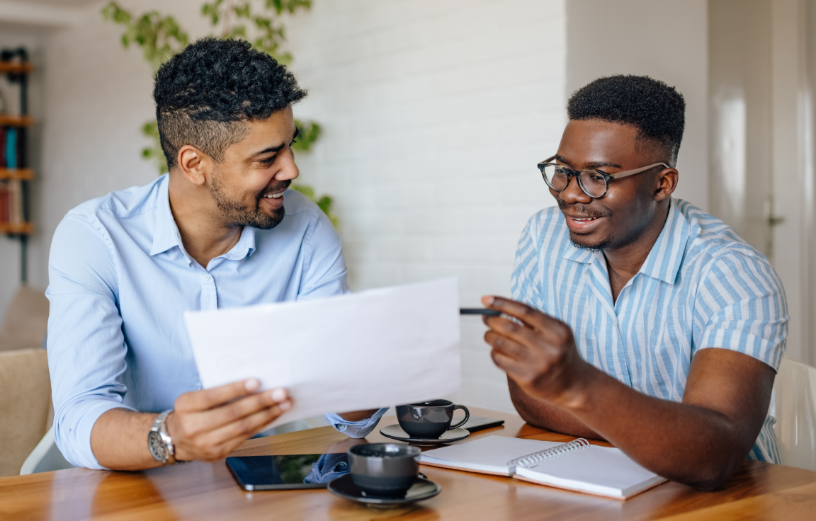 Two men sit at a table, smiling and discussing a document. One holds the paper while the other gestures with a pen. A notebook, tablet, and coffee cups are on the table. They appear engaged in a friendly conversation.