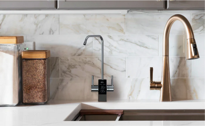 Modern kitchen countertop with a Brio digital water faucet next to a gold pull-down kitchen faucet. The backsplash is white marble tile, and glass storage jars with wooden lids containing sugar and beans are on the left side.