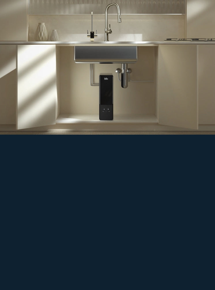 A black Brio Aquus under-sink water filtration system sits inside an open kitchen cabinet beneath a stainless steel sink. Above the counter, its smart faucet with a digital display stands beside the main kitchen faucet, in a warm, sunlit kitchen with cream cabinets.