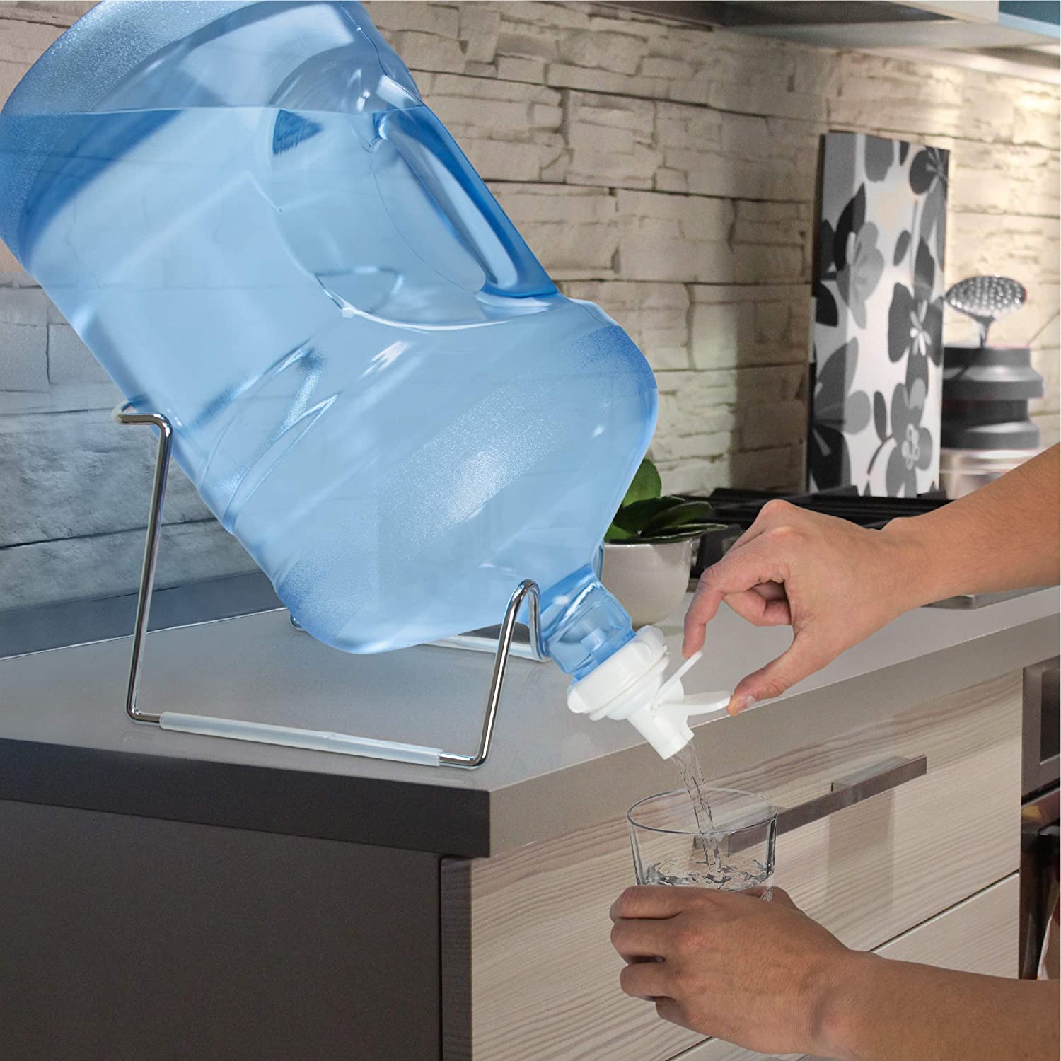 A person fills a glass with water from a large blue 5-gallon bottle placed on the Brio Metal-Plated Water Bottle Stand on the kitchen counter, with a stone wall and kitchen decor in the background.