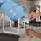 A person fills a clear glass with water from a 5-gallon blue bottle on the Stainless Steel Single Water Bottle Stand - White, placed next to a stainless steel sink on the kitchen counter.