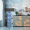 A modern kitchen features a rustic white and exposed brick wall, a wooden countertop with a sink, fruit bowl, potted plant, and the Single Column 4-Bottle Storage Rack - Gray holding four large blue water jugs.