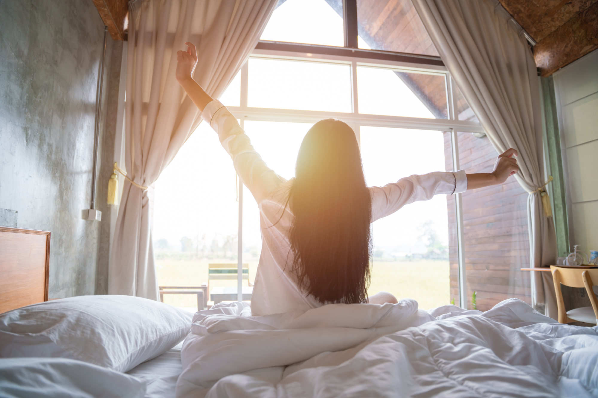 A woman waking up in the morning, stretching in the bed with the sun shining on her from a window
