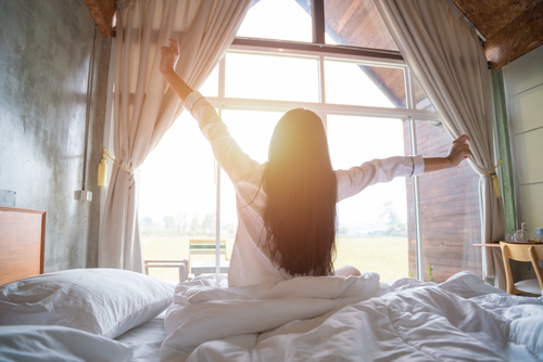 A woman waking up in the morning, stretching in the bed with the sun shining on her from a window