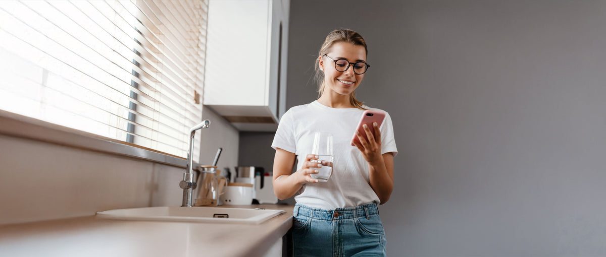 Woman in a kitchen holding a glass of water while checking her phone