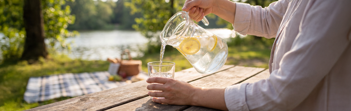 Pouring lemon-infused water from a pitcher into a glass at an outdoor picnic by a lake