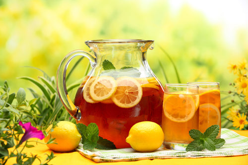 A glass pitcher filled with iced tea and lemon slices, flanked by whole lemons and two filled glasses of iced tea.