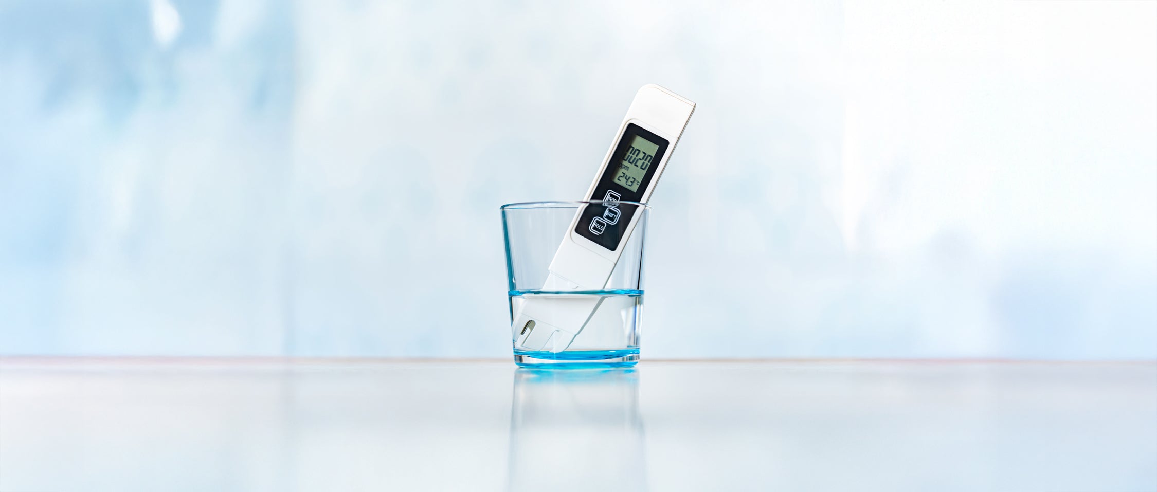 A digital TDS (total dissolved solids) meter is placed in a clear glass of water, with its display showing a reading. The background is softly blurred in light blue tones, giving the image a clean and minimal look.