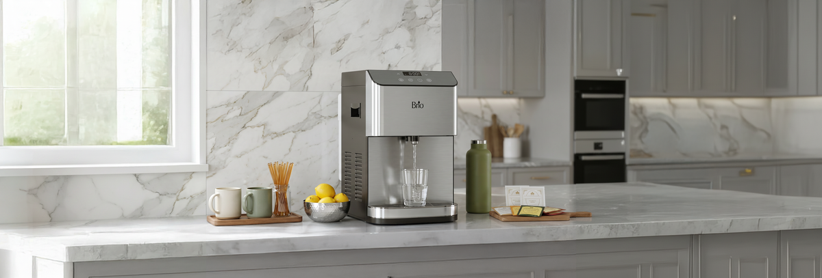 Stainless steel Brio Moderna countertop water dispenser pouring water into a glass on a modern marble kitchen counter, surrounded by mugs, lemons, and tea items.