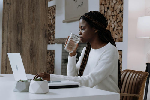 woman working on a laptop at a desk, sipping on a glass of water