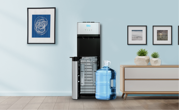 Brio bottom load filtered water cooler with a 5-gallon water bottle beside it, shown in a bright modern living room with light blue walls and framed art.