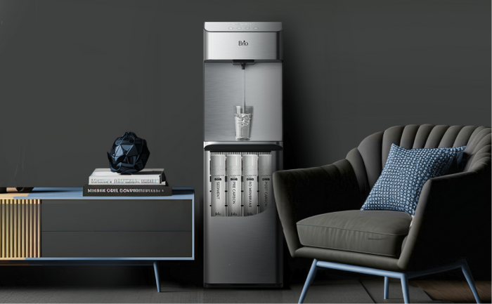 Brio stainless steel reverse osmosis water cooler with visible internal filters, displayed in a modern living space with a dark wall, accent chair, and side cabinet.