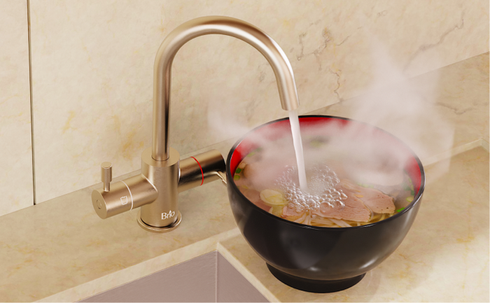 Brio hot water faucet in brushed gold finish pouring steaming water into a black bowl of ramen on a marble countertop.