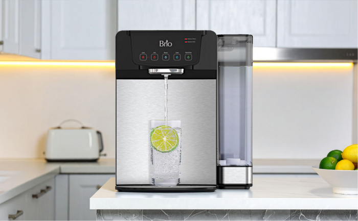 Brio countertop water dispenser on a kitchen counter, dispensing sparkling water with lime slices in a glass beside a bowl of fresh citrus fruit.