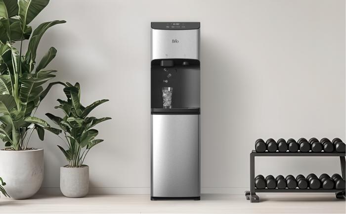 Brio stainless steel water and ice dispenser placed in a minimalist home gym setting with potted plants and a rack of dumbbells.