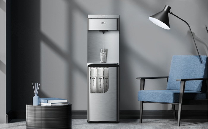 Brio bottleless water cooler with built-in filtration system, dispensing water in a stylish living room with a blue chair and floor lamp.