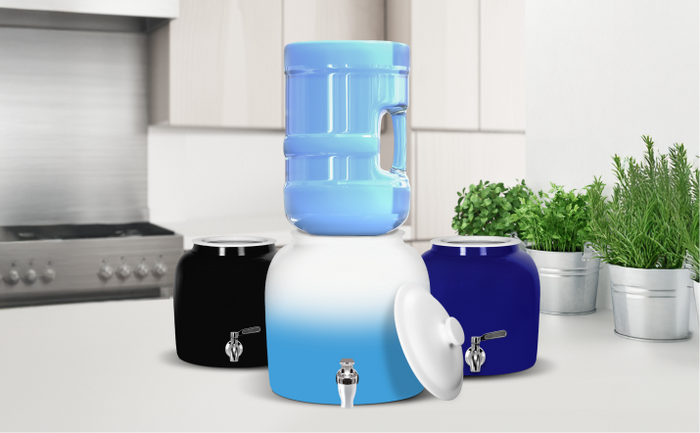 Brio ceramic water crock dispensers in black, blue, and ombre white, holding a 5-gallon water jug on a kitchen counter with fresh herbs.