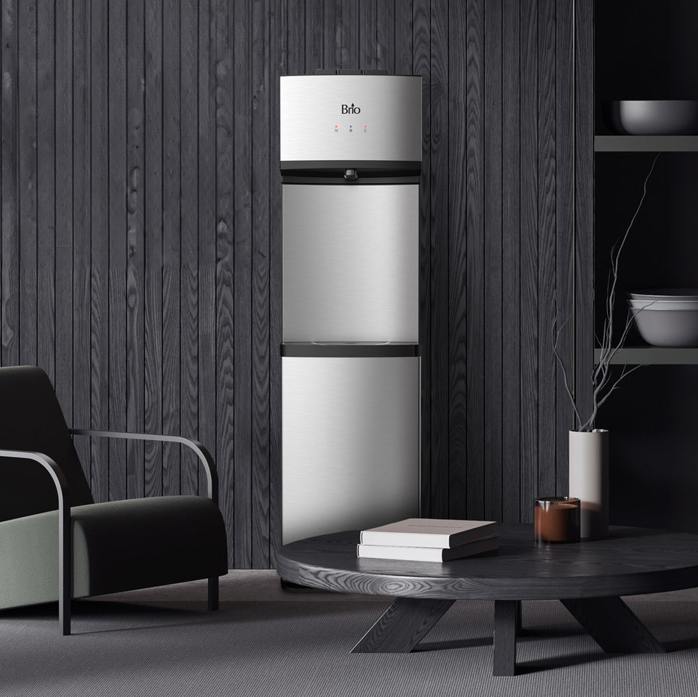 A stainless steel Brio 530 Series water dispenser stands against a dark wood-paneled wall in a moody, modern living space, next to a black armchair and a low round wooden coffee table topped with books, a candle, and a vase of bare branches.