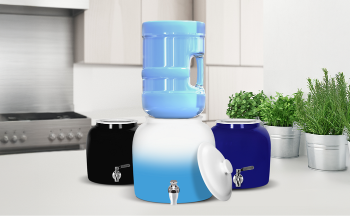 Brio ceramic water crock dispensers in blue, black, and white with a 5-gallon water bottle on top, displayed on a modern kitchen counter.