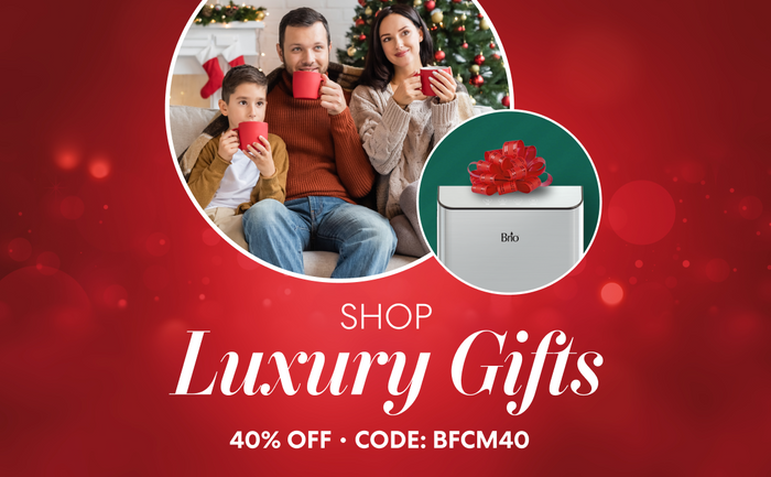 Holiday-themed Brio promotion with a red background. Text reads “Shop Luxury Gifts – 40% Off • Code: BFCM40.” The image shows a family of three sitting together in front of a Christmas tree and fireplace, holding red mugs, and a Brio water dispenser wrapped with a red bow.