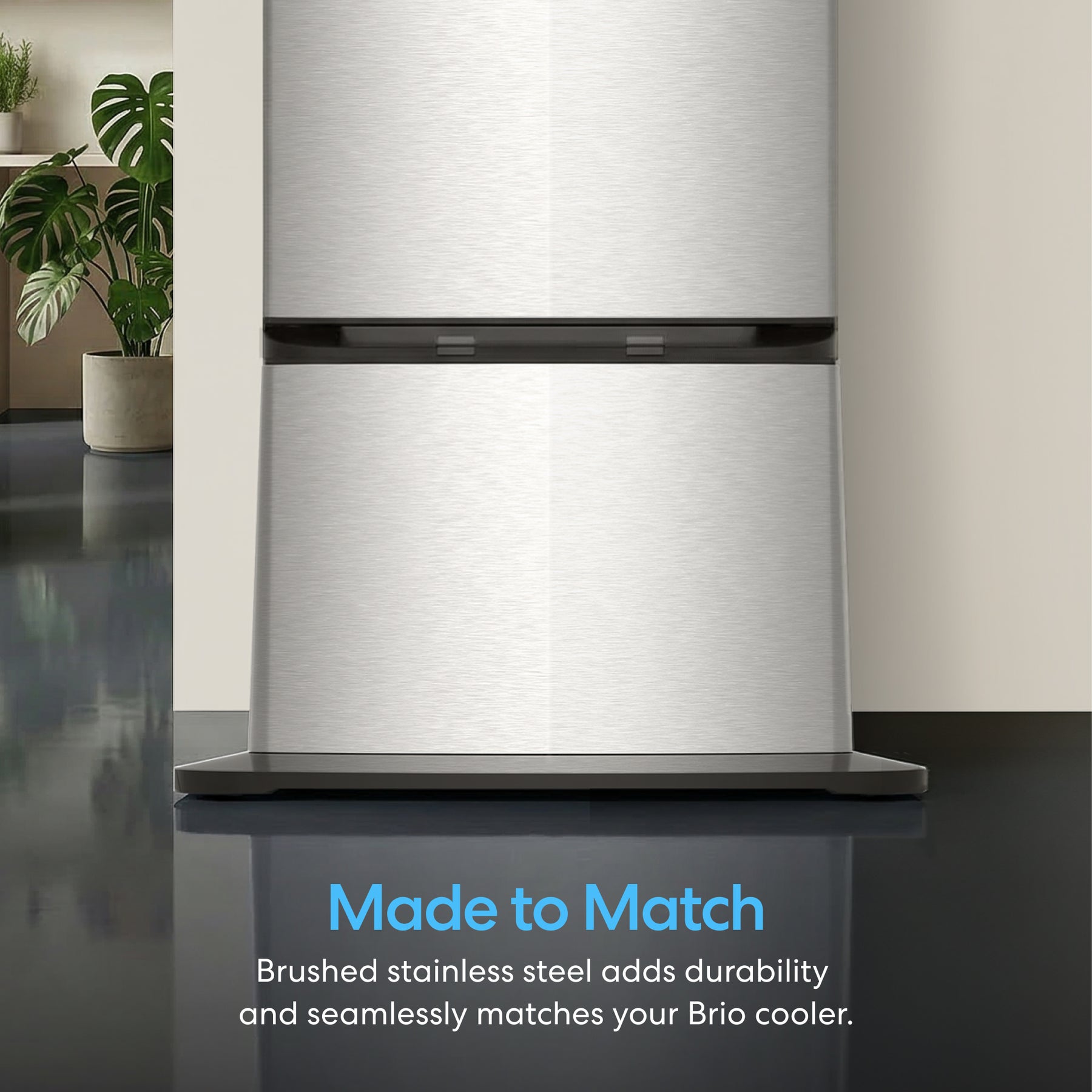 A close-up of the brushed stainless steel Brio 745 Series Compatible Cooler Base for Water Dispenser on a reflective floor with green plants behind it. Text: Made to Match. Durable brushed stainless steel complements your Brio water cooler.