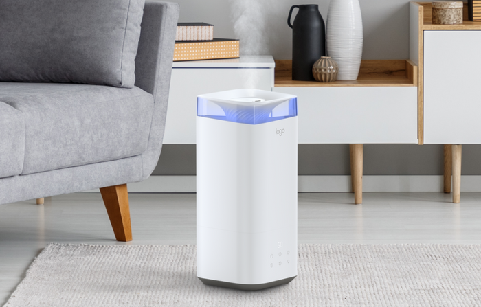 A white humidifier with a blue light on top emits mist in a modern living room, placed on a light wood floor near a gray sofa and a white cabinet with decorative items.
