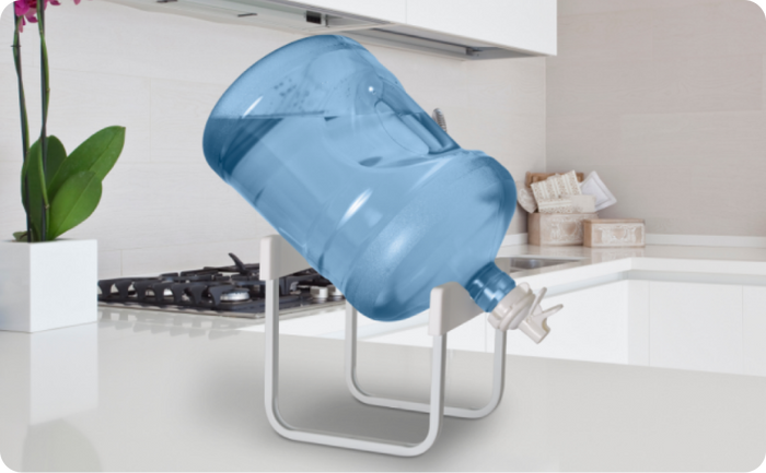 A large blue water jug is placed upside down on a white metal stand on a kitchen counter, with a white stove and a potted orchid visible in the background.