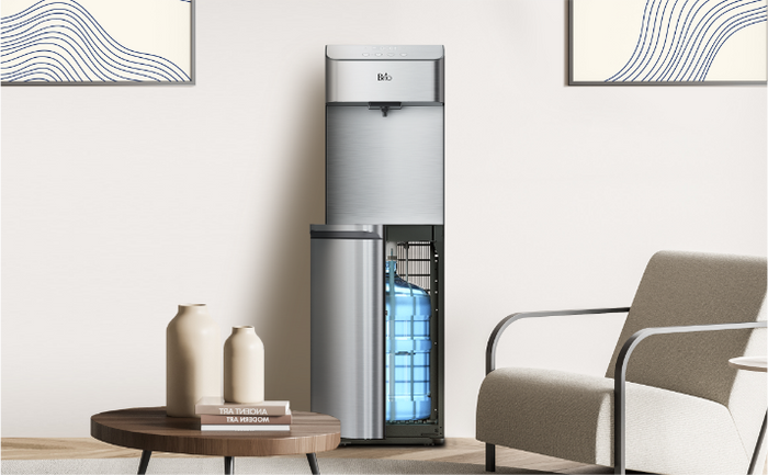 A modern water dispenser with a bottom-loading blue water bottle stands in a minimalist living room beside a beige armchair and a round wooden table with two vases. Abstract artwork hangs on the light-colored walls.