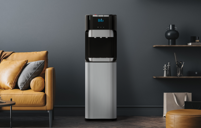A modern water dispenser stands in a stylish living room with a tan leather sofa, dark gray walls, decorative shelves, and minimal decor. The dispenser has a digital display and a metallic lower section.