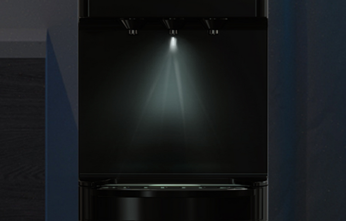 A close-up of a sleek, black appliance with a single bright light shining down from the top, illuminating an empty compartment below. The surrounding area is dark, highlighting the illuminated space.