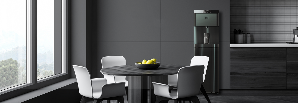 A modern kitchen with dark cabinets, a round table with four white chairs, a bowl of lemons on the table, a water dispenser, and large windows letting in natural light.
