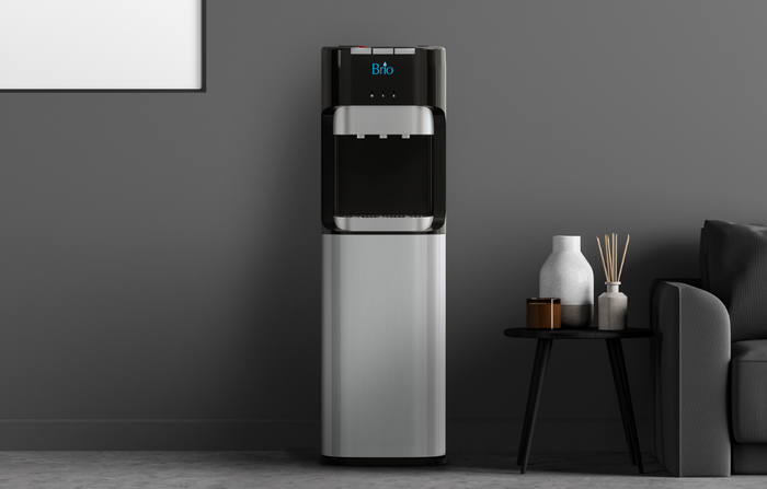 A modern water dispenser with a digital display stands against a gray wall next to a black round side table holding decorative vases and reed diffusers beside a dark sofa.