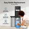 A person slides a large water bottle into the bottom compartment of the black Bottom Load Bullet Ice & Hot/Cold Water Dispenser – Model 420 Ice. Icons highlight easy bottle replacement, hassle-free use, and compatibility with 3- or 5-gallon bottles, while the UV self-cleaning feature ensures fresh and clean drinking water with every use.