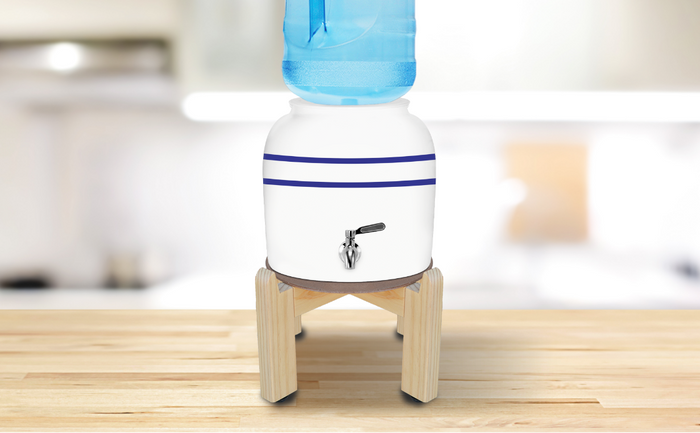 White ceramic water dispenser with two blue horizontal stripes and a silver spigot, placed on a light wooden stand, topped with a large blue water bottle, situated on a wooden countertop.