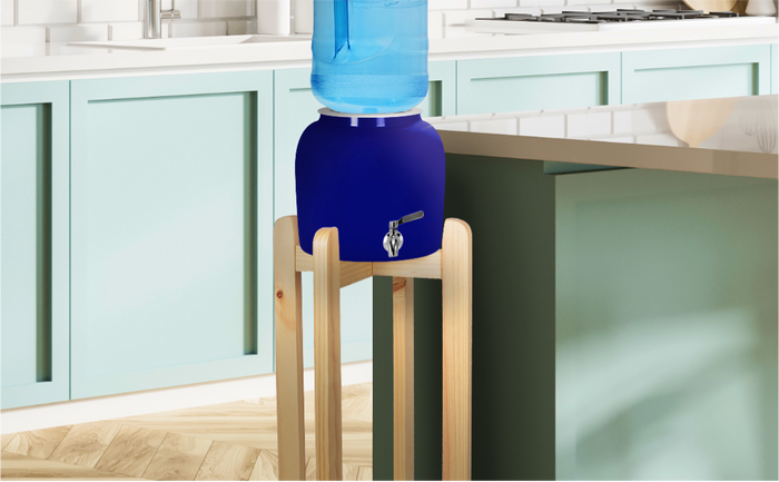 Blue ceramic water dispenser with a silver spigot, placed on a light wooden stand, topped with a large blue water bottle, situated in a kitchen with mint green cabinets and a white countertop.